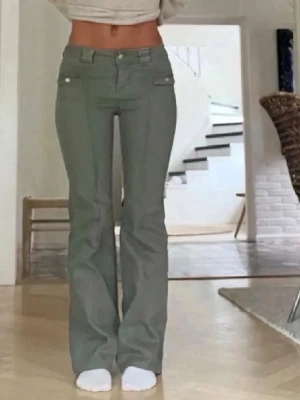 Olivgröna bootcut jeans med fickor - Säljer ett par olivgröna bootcut jeans med låg midja och dekorativa fickor framtill. Jeansen har vita kontrastsömmar och knappdetaljer. Säljer då dom är för korta för mig som är 178cm.