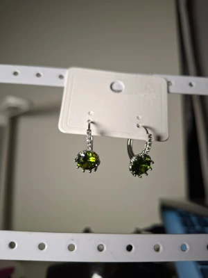 Silverörhängen med gröna stenar - Snygga örhängen i silverfärg med runda, gröna stenar i centrum och små klara stenar längs bågen. Perfekt för dig som gillar stilrena smycken med en färgklick. Materialet ser ut att vara metall med glittrande stenar.