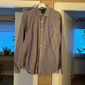Rutig skjorta Tommy Hilfiger L - Snygg rutig skjorta från Tommy Hilfiger i custom fit-modell. Skjortan har små rutor i rött, blått och vitt, klassisk krage, bröstficka och långa ärmar med knappar vid manschetterna. Perfekt för dig som gillar stilrena och tidlösa plagg.