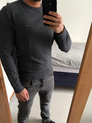Mörkgrå stickad tröja Tommy Hilfiger - Mörkgrå stickad tröja från Tommy Hilfiger med rund halsringning och diskret logga på bröstet i storlek S. Tröjan har långa ärmar och ribbade muddar vid ärmslut och nederkant. Perfekt för en stilren och enkel look och nu till våren! Inga defekter! Bara att kontakta vid funderingar!🌟