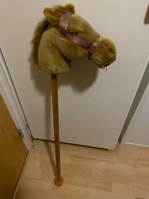 Käpphäst med brunt tyg och träpinne - Käpphäst med mjukt brunt tyg, bruna tygtyglar och broderade detaljer. Hästens huvud är stoppat och har en man av syntetmaterial. Pinnen är av trä och har en orange plastboll längst ner. Perfekt för lek och fantasi.