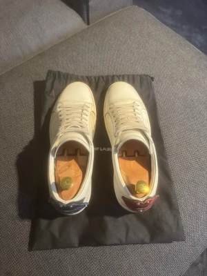 Saint Laurent vita sneakers med detaljer - Säljer ett par vita Saint Laurent sneakers i läder med klassisk låg profil. Skorna har kontrasterande hälflikar, en i rött och en i blått, samt vita snören och diskreta sömdetaljer. Perfekta för dig som gillar stilrena och exklusiva sneakers. Strl 43 men passar defenetivt 44