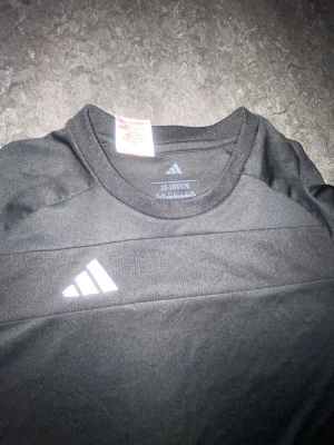 Svart sporttröja från Adidas - Snygg svart sporttröja från Adidas med klassisk vit logga på bröstet. Tröjan har rund halsringning och är tillverkad i ett lätt och funktionellt material som passar perfekt till träning eller sportaktiviteter.