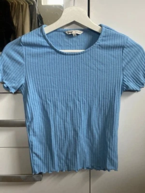 Blå ribbad topp från ONLY - Snygg ljusblå ribbad topp från ONLY med korta ärmar och rund halsringning. Toppen har en croppad passform och är tillverkad i mjuk bomullsmix som känns skön mot huden. Perfekt att matcha med jeans eller kjol för en fräsch look.