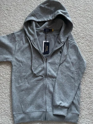 Grå hoodie från Polo Ralph Lauren - Snygg grå hoodie från Polo Ralph Lauren med dragkedja framtill, justerbar huva med snören och klassisk logga broderad på bröstet. Tillverkad i mjuk bomullsblandning och har känguruficka. Perfekt för en chill och stilren look. Självklart aldrig använd. Tveka inte på att höra av er om ni har frågor 😊👌🏻