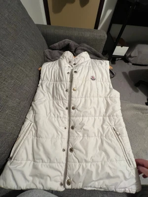 Vit dunväst från Moncler - Snygg vit dunväst från Moncler med hög krage och tryckknappar framtill. Västen har två dragkedjeförsedda fickor och det klassiska Moncler-logotypen på bröstet. Perfekt för lager-på-lager och riktigt clean look.