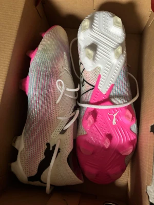 Puma fotbollsskor vit/rosa - Säljer ett par snygga fotbollsskor från Puma i vitt med rosa och svarta detaljer. Skorna har en modern design med stickad ovandel och fasta dobbar i plast, perfekta för spel på gräs. Lätta och smidiga med klassisk snörning och stor Puma-logga på sidan.