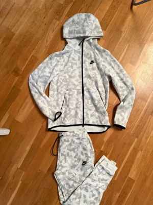 Nike ”Tech Fleece” set med grått camomönster - Nike hoodie och byxor i vitt och ljusgrått camomönster med svart dragkedja och svarta detaljer. Klassisk huva och två fickor framtill. Tillverkad i mjukt och stretchigt material som passar perfekt för en avslappnad stil. Nike-logga broderad.                Hoddie är i storlek S.                                                 Byxor är i storlek M                                                   Finns i Helsingborg. Kan även sälja byxor och hoddie separat. 