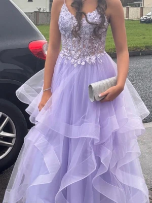 Balklänning - Säljer en lila långklänning med tunna axelband, spetsdetaljer och blommigt mönster upptill. Kjolen har flera lager av volang i tyll och snörning i ryggen. Perfekt för dig som vill sticka ut med en romantisk look.