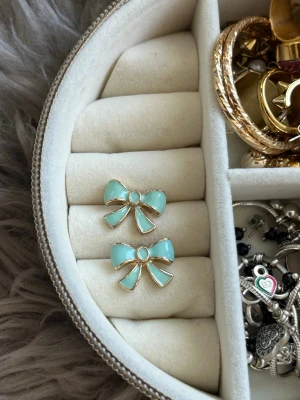 Turkosa rosettörhängen i emalj - Säljer ett par söta örhängen formade som rosetter i turkos emalj med guldfärgade kanter. De har en lekfull och trendig design som verkligen sticker ut. Perfekta för dig som vill addera en färgklick till din stil.