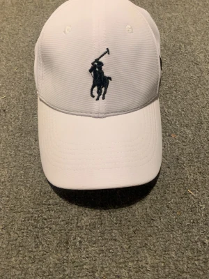 Vit keps från Polo Ralph Lauren - Vit keps med klassisk Polo Ralph Lauren-logga broderad framtill och en svart siffra 3 på sidan. Kepsen har böjd skärm och justerbar passform. Perfekt accessoar för en sportig och stilren look.