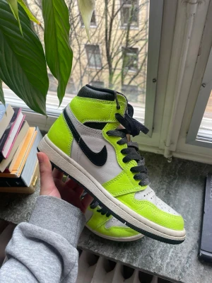 Nike Air Jordan 1 High Volt Neon - Säljer ett par Nike Air Jordan 1 High sneakers i färgen volt/neon, vitt och svart. Skorna har klassisk hög siluett, svart swoosh, svarta skosnören och detaljer i neonfärg. Ovandelen är i läder och mesh för en fräsch och sportig vibe.