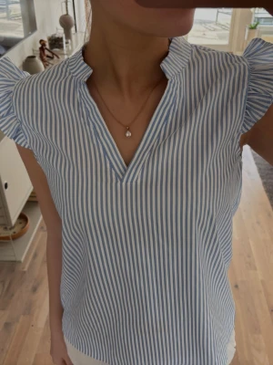 Blåvit randig blus med volangärm - Snygg blus med blå och vita vertikala ränder, v-ringning och volangdetaljer vid axlarna. Perfekt för en fräsch och somrig look. Storlek xs-s 