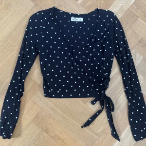 Svart omlottopp med prickar från Hollister - Svart omlottopp från Hollister, perfekt långärmad festtopp!! Toppen har knyt i sidan och v-ringning framtill. Köpt på Sellpy knappt använt själv. 