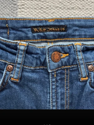 Blå skinny jeans från Nudie Jeans - Snygga blå skinny jeans från Nudie Jeans med klassisk femficksdesign och orangea kontrastsömmar. Jeansen har hög midja och är tillverkade i denim med stretch för en bekväm passform. Perfekta för en trendig och avslappnad look.