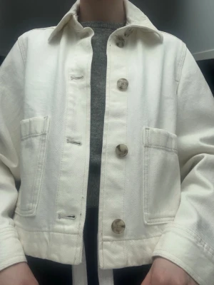 Vit jeansjacka med stora fickor - Vit jeansjacka med stora fickor framtill. Den stängs med fyra stora knappar. Lite för stor och tung för min smak. Den ser tunn ut men är ganska tjock i materialet och tycker inte att passformen passar på mig. Använd en gång. Den har en ”smutsvit”/beige färg. 
