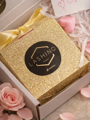 Lashino guldglittrig fransbox - Lyxig guldglittrig förpackning från Lashino, perfekt för lösögonfransar. Boxen har en elegant svart logga och stängs med en gul sidenrosett. Snygg och trendig design som gör sig fin på sminkbordet.