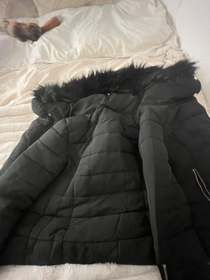 Svart dunjacka med fuskpäls - Snygg svart dunjacka med quiltat mönster och huva med svart fuskpäls. Jackan har dragkedja i guld och praktiska fickor. Perfekt för kalla vinterdagar och ger en trendig look. Materialet är vadderat och håller dig varm hela säsongen.