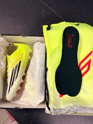 Adidas F50 gula fotbollsskor - Säljer ett par Adidas F50 fotbollsskor i neon-gult med svarta och röda detaljer. Skorna har klassisk låg profil, snörning och dobbar för spel på gräs. Medföljer matchande skopåse och extra innersulor. Perfekta för dig som vill sticka ut på planen. Vid snabb affär kan priset sänkas