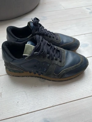 Navy Valentino Rockrunners - Snygga marinblå sneakers från Valentino med coola nitar på sidorna. Skorna har en mix av mocka och läder, mörkblå skosnören och en brun sula. Perfekta för dig som gillar grisch stilen och vill ha något unikt på fötterna. Skorna är en size 40 men sitter som en storlek 41a. De är skavda bak på sulan och saknar 3 spikar på sidorna. Det är bara att skriva om det skulle vara något du funderar över🕺