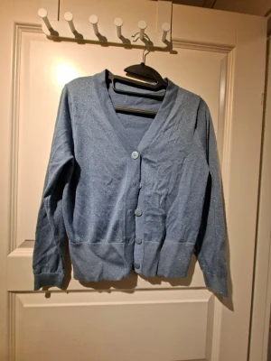 Blå glittrig kofta med v-ringning - Snygg blå kofta med diskret glittrigt material och v-ringad hals. Koftan har långa ärmar, bred mudd i nederkant och fyra knappar framtill. Perfekt att slänga över en t-shirt för en chill men ändå stylish look.