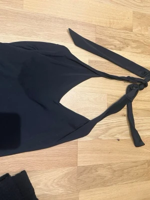 Svart halterneck linnetopp - Svart linnetopp i halterneck-modell med breda band som knyts i nacken. Toppen är ärmlös och har en djup urringning framtill. Materialet känns stretchigt och mjukt, troligtvis polyester eller syntet.