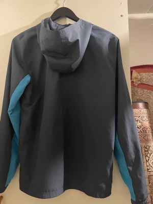 Blå vindjacka från Under Armour - Snygg marinblå vindjacka från Under Armour med ljusblå detaljer längs ärmarna. Jackan har huva, dragkedja framtill och två sidofickor. Perfekt för blåsiga dagar och sportiga aktiviteter. Tillverkad i lätt och vindtätt material.