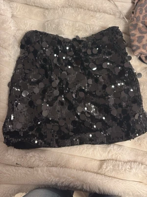 Svart paljettkjol med stora paljetter - Supersnygg svart kortkjol täckt med stora glittriga paljetter som verkligen fångar ljuset. Perfekt för dig som vill sticka ut och älskar att glänsa. Kjolen har en rak passform och är lätt att matcha med olika toppar. 