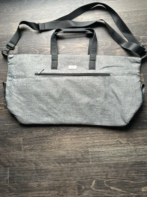 Grå axelväska från BOSS - Snygg grå axelväska från BOSS med svart justerbar axelrem och två handtag. Väskan har en stor ytterficka med dragkedja och en metallbricka med BOSS-logga på framsidan. Tillverkad i slitstarkt tygmaterial med stilren design.