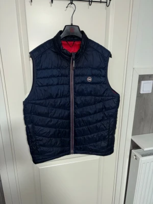 Mörkblå Väst från Jack & Jones - Snygg mörkblå dunväst från Jack & Jones med quiltad design och dragkedja framtill. Västen har röda och vita detaljer längs dragkedjan och ett diskret logomärke på bröstet. Perfekt att slänga över en hoodie eller tröja för extra värme.