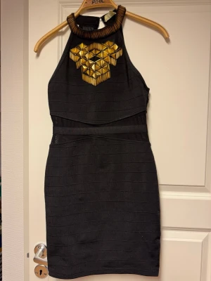 Svart kort klänning med gulddekor - Snygg svart kort klänning  med hög hals och unika guldfärgade geometriska dekorationer framtill. Klänningen har en kroppsnära passform och är ärmlös, perfekt för dig som vill sticka ut med coola detaljer.