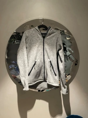 Grå hoodie med dragkedja från Everest - Säljer en gråmelerad hoodie från Everest med hel dragkedja framtill och två snedställda fickor med svarta dragkedjor. Hoodien har huva och är tillverkad i ett mjukt, fleeceliknande material som känns riktigt skönt mot huden. Perfekt för chill dagar eller när du vill ha något bekvämt.