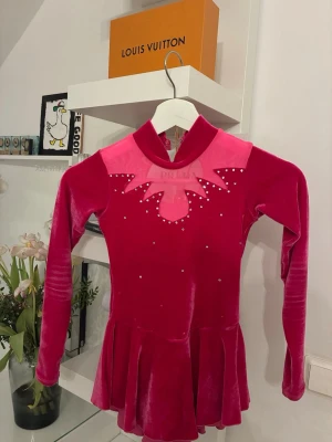 Rosa konståkningsklänning med strass - Snygg konståkningsklänning från Prima i mörkrosa sammet med ljusrosa meshdetaljer och glittrande strass på bröstet och ryggen. Klänningen har lång ärm, hög krage och en rund öppning i ryggen. Kjoldelen är veckad för extra rörelsefrihet på isen.