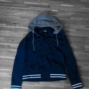 Blå vindjacka med huva från Hollister - Snygg marinblå vindjacka från Hollister med grå huva och vita ränder på muddar och nederkant. Jackan har knappar framtill och dragsko i huvan. Perfekt för dig som vill ha en sportig och avslappnad stil.
