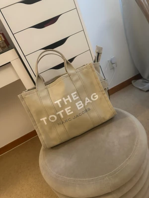 Beige The Tote Bag från Marc Jacobs - Snygg beige shoppingväska från Marc Jacobs med texten 'THE TOTE BAG' tryckt i vitt på framsidan. Väskan är tillverkad i kraftigt canvasmaterial och har dubbla handtag för enkel bärning. Perfekt för att bära med sig allt man behöver under dagen. Har användts sparsamt, men har en defekt som är en sminkfläck på kortsidan. Men upplever inte att detta är något man ser supertydligt. Inköpt för runt 3000kr, släpper för 1000kr.