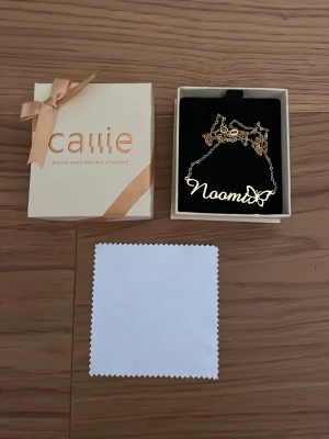 Guldigt namnhalsband med fjäril - Personligt halsband i guldfärgad metall från Callie med namnet 'Noomi' och en söt fjärilsdetalj. Kedjan är tunn och elegant, och halsbandet levereras i en fin presentask med rosett. Perfekt accessoar för att lyfta din stil. Helt ny, alldrig använd