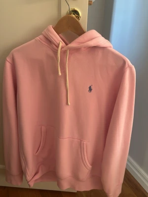 Rosa hoodie från Polo Ralph Lauren - Snygg rosa hoodie från Polo Ralph Lauren med klassisk huva och dragsnören. Har en broderad logga i blått på bröstet och en stor magficka framtill. Perfekt för en chill och avslappnad stil.