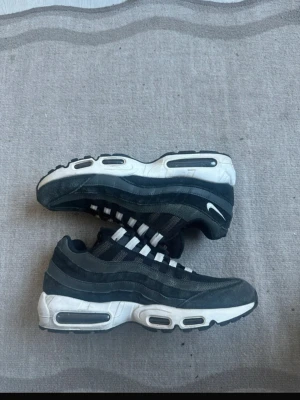 Svarta Nike Air Max 95 sneakers - Säljer ett par svarta Nike Air Max 95 sneakers med vit sula och synliga Air-enheter. Skorna har en vågig design i flera nyanser av svart och grått, samt vita detaljer på snörningen och sulan. Klassisk Swoosh-logga på sidan och mesh- samt mockaliknande material.