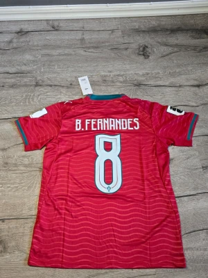 Portugal matchtröja B. Fernandes #8 - Snygg röd Portugal matchtröja från Puma med nummer 8 och B. Fernandes på ryggen. Tröjan har vita detaljer, vågmönster och gröna inslag vid krage och ärmslut. Perfekt för dig som älskar fotboll och vill stötta landslaget.