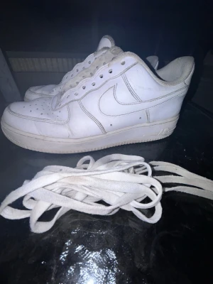 Vita Nike Air Force 1 - Klassiska vita Nike Air Force 1 sneakers i lågt utförande. Skorna är tillverkade i läder med perforerade detaljer på tån och har den ikoniska swoosh-loggan på sidan. Medföljer vita skosnören. Perfekta för en clean och tidlös streetstil. Skorna är använda och har tydliga tecken på användning, vilket syns på bilderna. Trots det finns det fortfarande mycket användning kvar och de passar perfekt som vardagsskor eller ett prisvärt par Air Force 1. Storlek 42.5