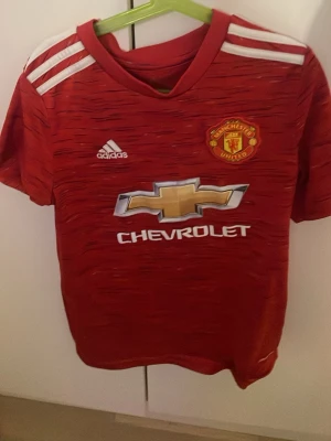 Manchester United matchtröja röd - Röd Manchester United matchtröja från Adidas med klubbmärke och sponsortryck på bröstet. Tröjan har vita detaljer på axlarna och klassisk rund hals. Perfekt för fotbollsträning eller att bära på matchdagar.