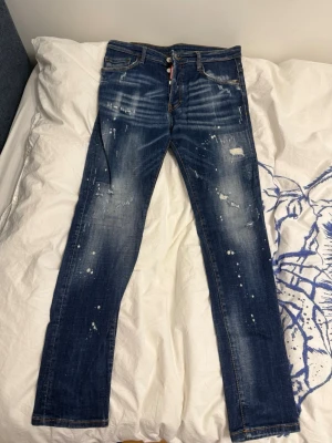 Mörkblå distressed jeans Dsquared2 - Säljer ett par mörkblå jeans från Dsquared2 med slitningar och färgstänk för en trendig, avslappnad look. Jeansen har raka ben, klassiska fem fickor och kontrastsömmar. Perfekta för dig som gillar streetwear och vill sticka ut.
