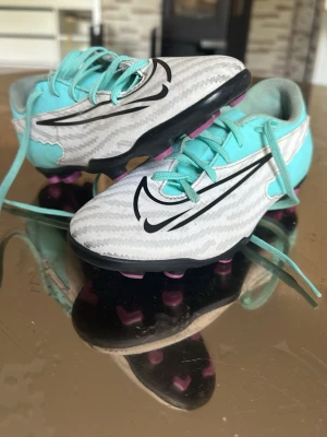Nike turkosa och vita fotbollsskor - Säljer ett par snygga fotbollsskor från Nike i turkos och vitt med svarta detaljer och stora Nike-loggor på sidorna. Skorna har snörning, mönstrad ovandel och dobbar i svart och lila för bra grepp på planen. Perfekta för dig som vill sticka ut på fotbollsplanen! Storlek 32