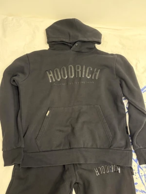 Svart tracksuit från Hoodrich - Svart tracksuit från Hoodrich med stor logga på bröstet och känguruficka framtill. Klassisk huva med knappdetalj och ribbade muddar vid ärmar och nederkant. Perfekt för en streetwear-look och riktigt skön att bära.