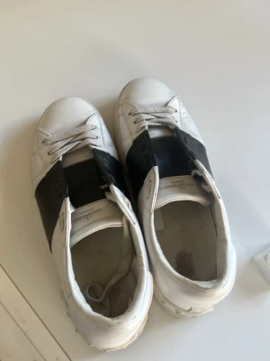 Valentino Garavani Sneakers - Stilrena vita sneakers med bred svart rem över snörningen. Klassisk låg modell med rund tå och vita skosnören. Tillverkade i slätt material som ger en clean look. Perfekta för dig som gillar minimalistisk stil och vill ha något som sticker ut lite extra.