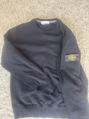 Svart sweatshirt från Stone Island - Svart sweatshirt från Stone Island med klassisk rund hals och ikonisk logotyp-patch på vänster ärm. Tröjan har ribbade muddar vid ärmslut och nederkant. Perfekt för en stilren och avslappnad look. Snygg stone island tröja som var väldigt dyr och fin