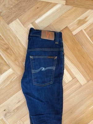 Mörkblå jeans från Nudie Jeans - Snygga mörkblå jeans från Nudie Jeans med klassisk femficksdesign och kontrastsömmar. Jeansen har rak passform och är tillverkade i denim med en tydlig vågdetalj på bakfickan samt läderpatch i midjan.
