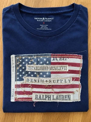 Denim&supply T-shirt - Snygg marinblå t-shirt från Ralph Lauren Denim & Supply med ett stort patch framtill i amerikansk flaggdesign och slitna detaljer. T-shirten har rund hals och korta ärmar, tillverkad i mjuk bomull för skön känsla.