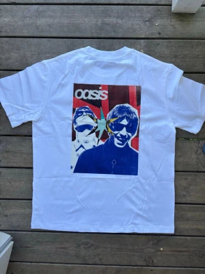 Vit Oasis t-shirt med tryck live forever - Vit t-shirt med stort färgglatt tryck på ryggen föreställande två personer och texten 'Oasis'. T-shirten har klassisk passform och korta ärmar. Perfekt för dig som gillar bandmerch och vill sticka ut med en unik design.