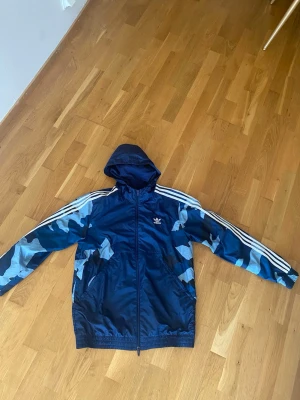 Blå camo vindjacka från Adidas - Snygg blå vindjacka från Adidas med camo-mönstrade ärmar i olika blå nyanser. Jackan har huva, dragkedja framtill och klassiska vita ränder på axlarna. Perfekt för dig som vill ha en sportig och trendig look. Materialet är lätt och vindtätt.
